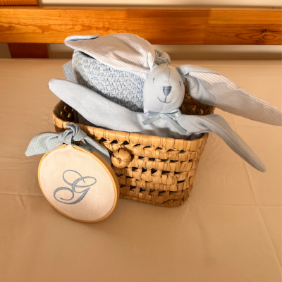 Cesto de verga com manta cinzenta e coelho de peluche azul claro com orelhas riscas e ornamento de madeira com letra G