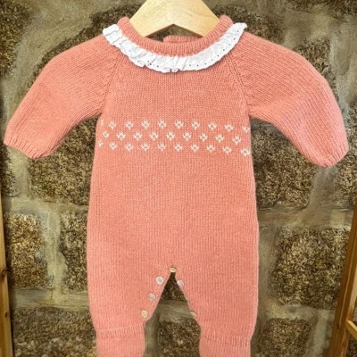 Macacão cor de rosa tricotado para bebé com gola branca bordada, em cabide de madeira contra parede de pedra