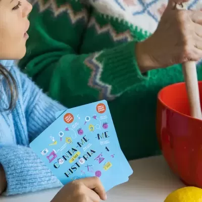 Criança segura cartas de jogo com texto, adulto mistura numa taça vermelha