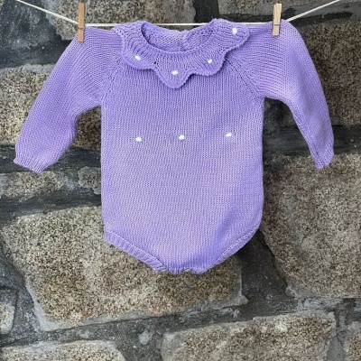 Body de bebé lilás em malha com gola ondulada e pompons, pendurado num estendal frente a parede de pedra.