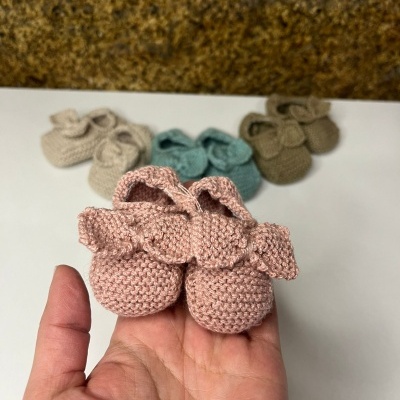 Botinhas de tricô para bebé em várias cores com laços, sobre superfície branca e parede texturada ao fundo.