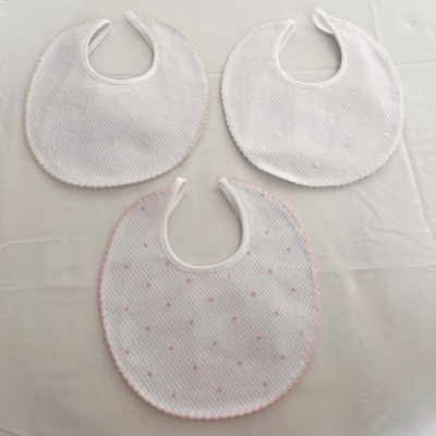 Três babetes de bebé redondos em tecido branco e com detalhes cor-de-rosa, sobre fundo branco.