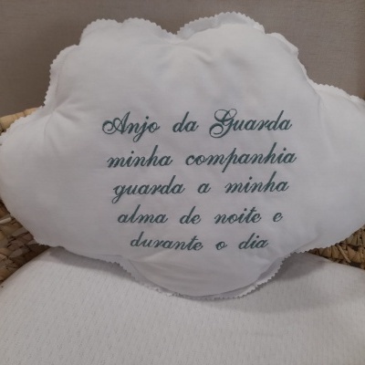 Travesseiro em forma de nuvem branco com bordado azul com texto em português, sobre superfície branca e cesta de palha.