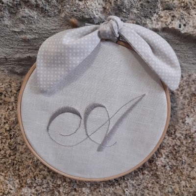 Bastidor de madeira com tecido branco bordado com a letra A e laço branco com pequenos pontos
