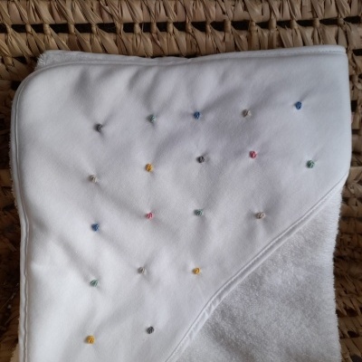 Toalha branca de bebé com capuz e bordados coloridos sobre superfície de fibras entrelaçadas