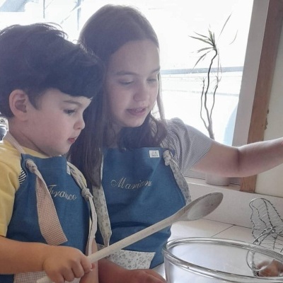 Duas crianças com aventais azuis personalizados a cozinhar perto de uma janela com luz natural