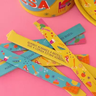 Tiras de papel coloridas com frases e ilustrações sobre superfície rosa próximas a latas amarelas