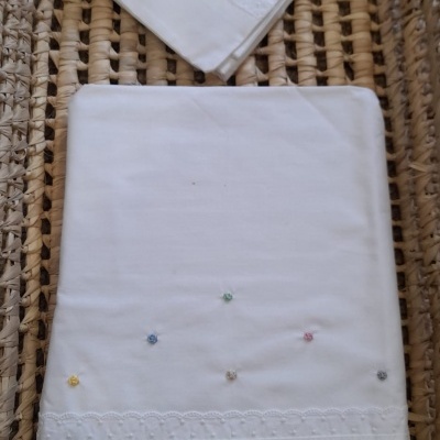 Conjunto de roupa de cama branca com bordados coloridos e detalhes em renda sobre superfície de palha trançada