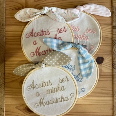 Bastidores de bordado com a frase 'Aceitas ser a minha Madrinha?' e laços decorativos em diferentes padrões e cores sobre madeira clara