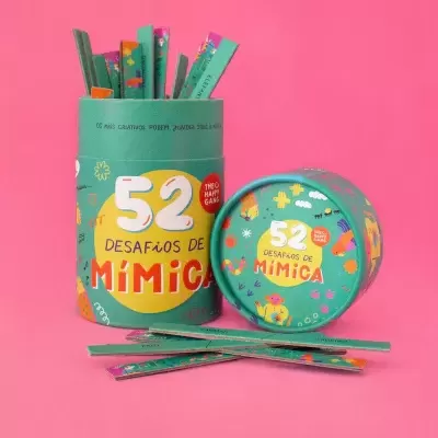 Jogo '52 Desafios de Mímica' com embalagem verde e pauzinhos de papel, sobre fundo rosa.