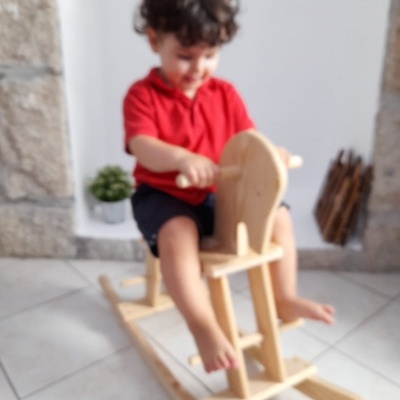 Menino sentado numa cavalo de madeira em chão de azulejos brancos junto a parede de pedra