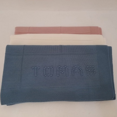 Três mantas de malha azul, branca e rosa empilhadas, manta azul com a palavra TOMAS em relevo