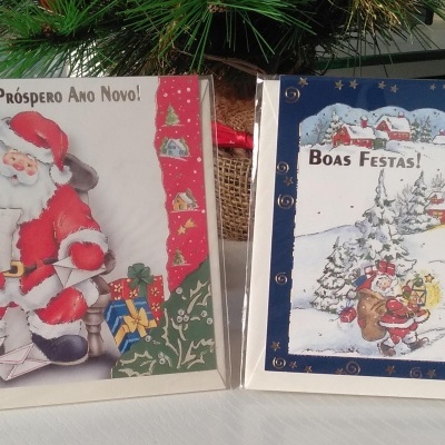 Dois cartões de Natal com imagens de Pai Natal e aldeia nevada com boneco de neve e textos festivos.