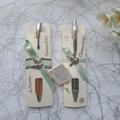 Dois conjuntos de canetas decoradas para comunhão com fitas verdes e pendentes de pomba em embalagem de cartão.