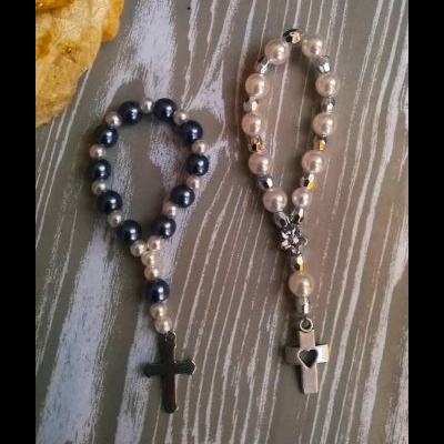 Dois terços em pérolas com crucifixos, um preto e outro prateado, sobre superfície cinza claro