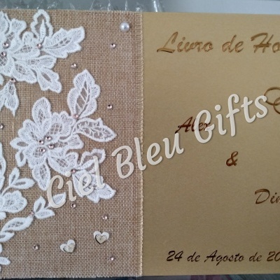 Livro de honra com capa bege, texto dourado e decoração de renda branca sobre juta