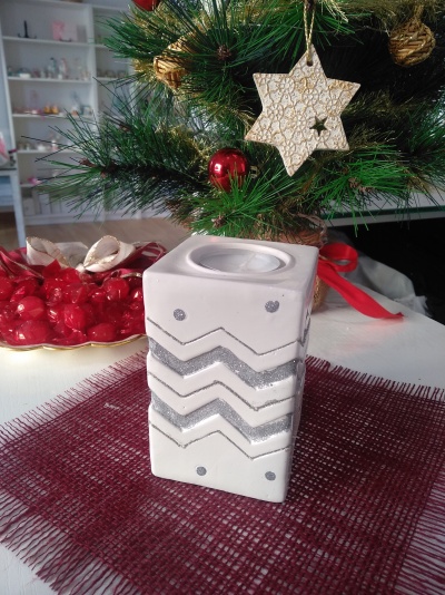 Vela decorativa quadrada branca com padrões prateados sobre tecido vermelho, junto a árvore de Natal com enfeites dourados e vermelhos.