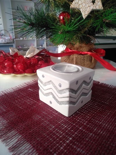 Suporte para vela branco com padrões prateados em tecido vermelho junto a árvore de Natal