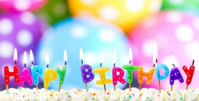 Velas coloridas 'HAPPY BIRTHDAY' acesas sobre bolo branco com balões coloridos ao fundo