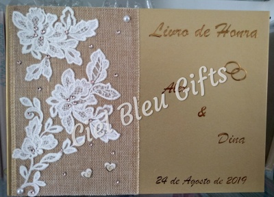 Livro de honra com capa bege, texto dourado e decoração de renda branca sobre juta