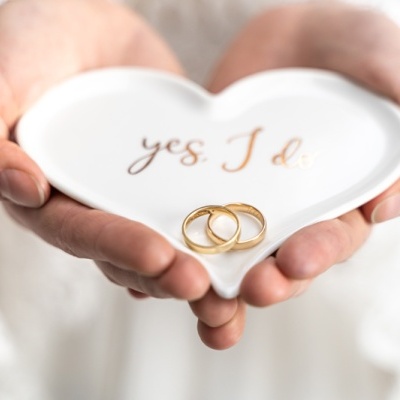 Prato branco em forma de coração com texto dourado e dois anéis de ouro sendo segurado por mãos