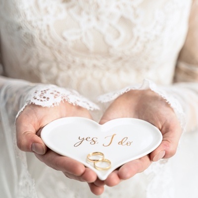 Prato coração branco com texto 'yes, I do' e dois anéis dourados em mãos com roupa de renda branca