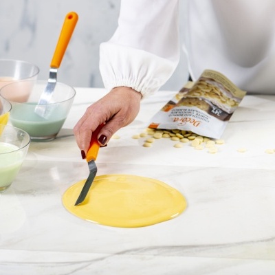 Pessoa mexendo massa amarela circular com espátula laranja, tigelas com líquidos coloridos e embalagem branca com tabletes amarelos ao redor