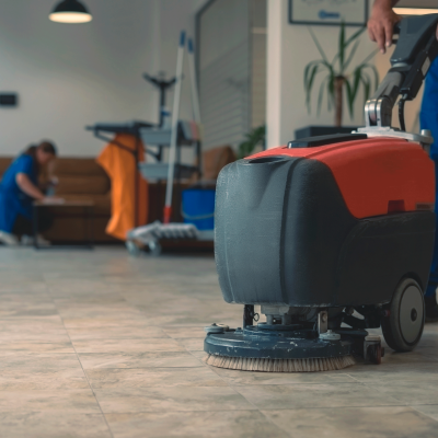 Máquina de limpeza de pisos vermelha e preta, operada por pessoa com roupa azul em interior com chão de azulejo cinzento