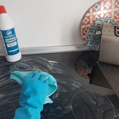 Mão com luva azul a limpar placa vitrocerâmica preta com produto de limpeza OTROHOG e pano verde na cozinha