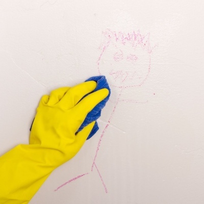 Mão com luva amarela a limpar desenho rosa numa parede branca com pano azul