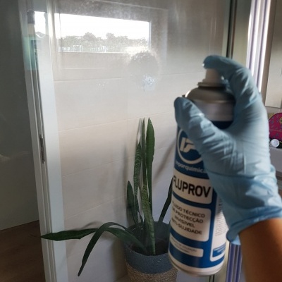 Mão com luva azul a segurar spray aerosol CIPROV junto a planta verde em vaso junto a parede branca