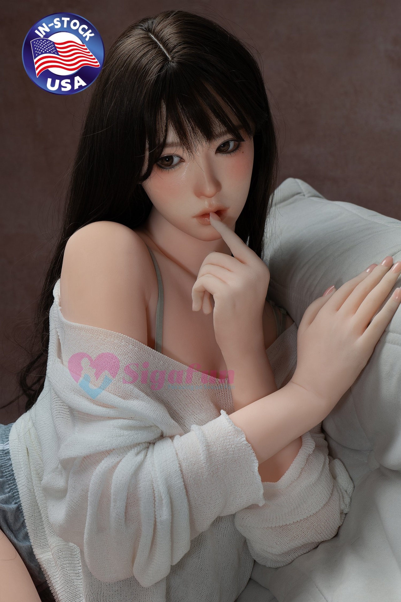 Boneca realista com cabelo castanho escuro e roupa branca sentada num sofá cinza claro