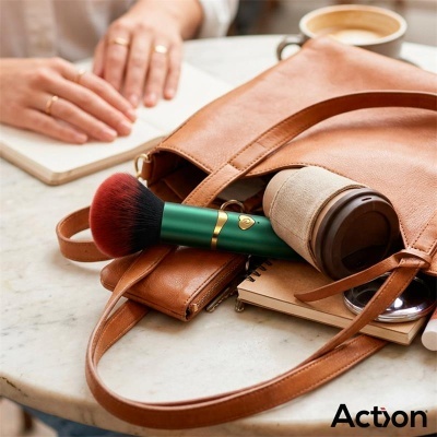 Bolsa de couro castanho com pincel de maquilhagem verde, garrafa térmica, caderno e estojo sobre mesa clara