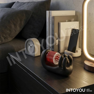 Mesa de cabeceira com relógio cinza, livros, suporte de telemóvel e luminária ao lado de um sofá cinzento