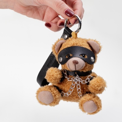Chaveiro urso peluche com máscara preta e corrente metálica na mão de pessoa com unhas vermelhas
