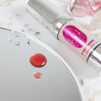 Frasco de clitoral balm com rótulo rosa ao lado de espelho com gotas vermelhas e cristais.