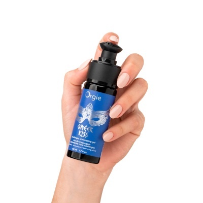Frasco de gel estimulante anal Orgie Greek Kiss segurado por mão feminina com unhas nude.