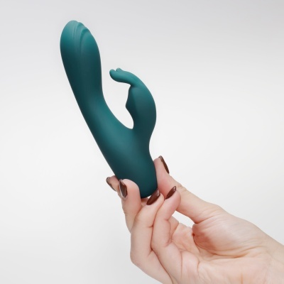 Vibrador de silicone verde escuro com duas extremidades curvas em fundo branco, segurado por mão.