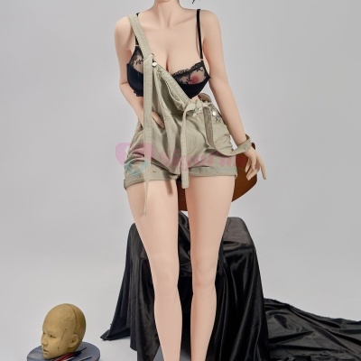 Boneca realista com roupa bege e soutien preto, em estúdio com acessórios de pintura no chão.