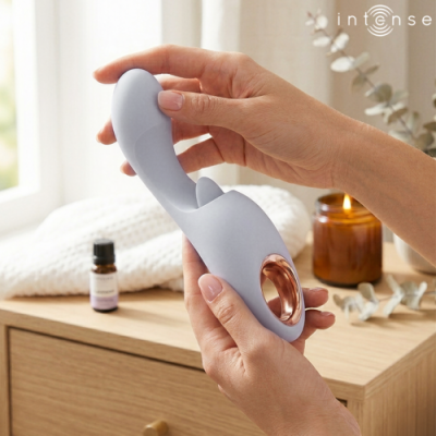 Vibrador cinza com detalhe dourado segurado por mãos, sobre mesa com óleo essencial, vela acesa e texto intense