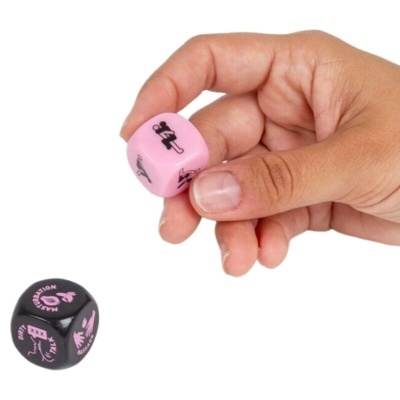 Dois dados pequenos coloridos, um rosa com ícones e outro preto com texto, segurados por uma mão sobre fundo branco.