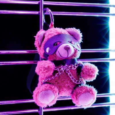 Urso de peluche cor de rosa com máscara, coleira e correntes metálicas.