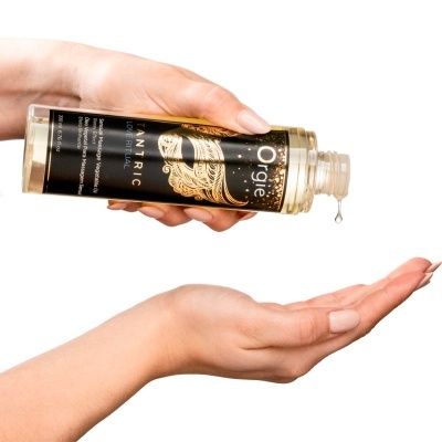 Frasco transparente de óleo para massagem com rótulo preto e dourado, óleo a cair na mão.