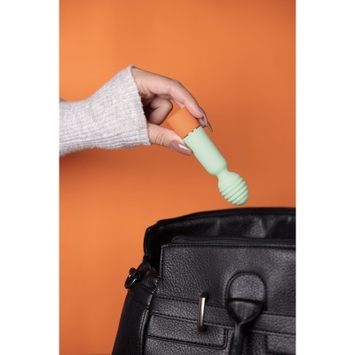 Mão segurando vibrador verde e laranja ao lado de mala preta em fundo laranja