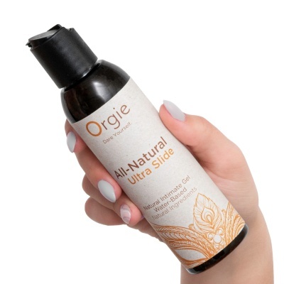 Frasco de gel íntimo Orgie All-Natural Ultra Slide com rótulo branco e texto laranja e preto, segurado por mão com unhas cinza