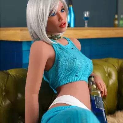 Boneca realística com roupa azul turquesa e cabelo branco sentada num sofá verde, segurando uma garrafa azul