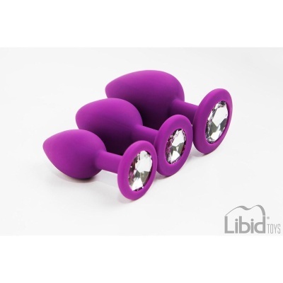 Três plugs anais roxos com pedras brilhantes em base, dispostos sobre fundo branco.