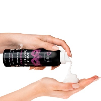 Frasco preto de espuma de massagem Orgie com tampo branco e espuma branca a sair, segurado por mãos femininas