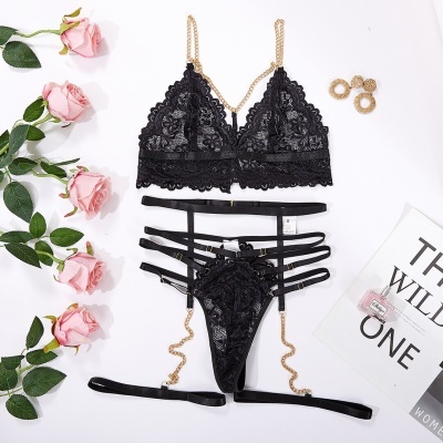 Conjunto de lingerie preto em renda com alças de corrente dourada sobre fundo branco com flores e acessórios.