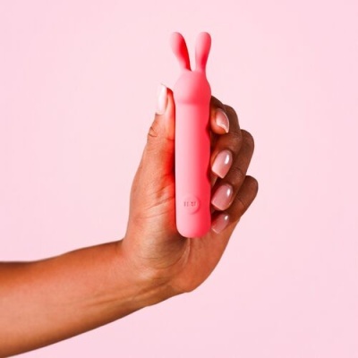 Mão segurando vibrador rosa com duas orelhas em fundo rosa